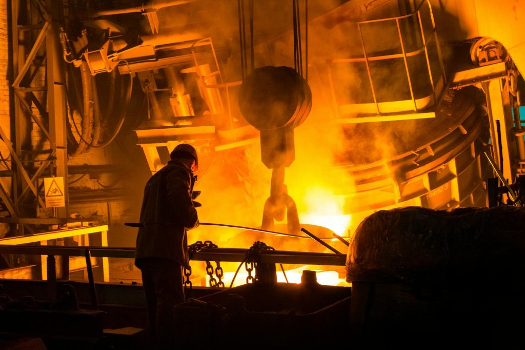 A steelworker in protective gear watches glowing molten iron and sparks beside a converter furnace in a steel mill
