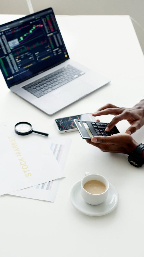 A person at a desk looking anxiously at a smartphone showing stock chat messages, with trading charts on a laptop