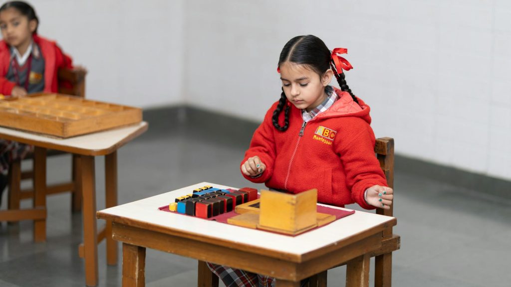A small child sits at a desk piled with textbooks and worksheets in a classroom