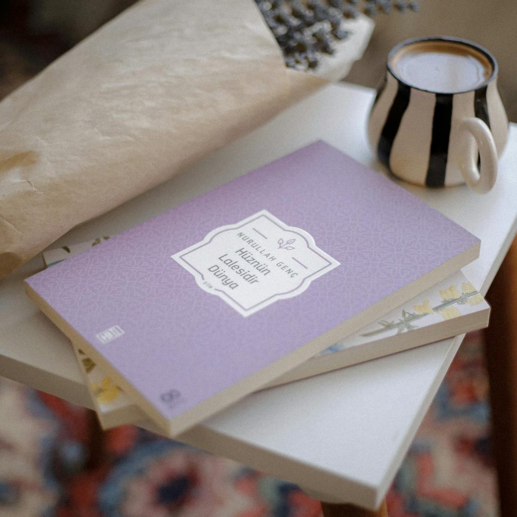 A stack of three books on a wooden table beside a teacup, with a newspaper blurred in the background